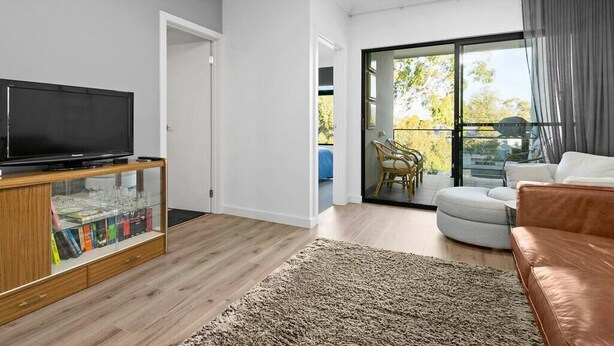 Ein modernes Wohnzimmer mit einem Flachbildfernseher auf einer Holzkommode, einem großen Fenster mit Blick auf Bäume und einem braunen Ledersofa.