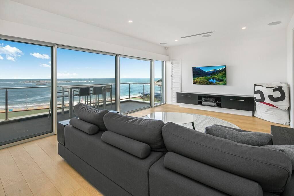 A modern living room with a large sofa, a flat-screen TV, and a balcony with a view of the sea.