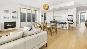 A modern kitchen with white cabinets, a central island with bar stools, and a dining area with a light-colored sofa and a fireplace. - Strangeways Stays! -Slps 10, 4 Bdrm, Oceanview (Port Elliot)