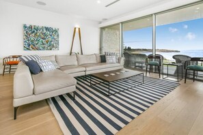 Un salon moderne avec un canapé beige, un tapis à rayures et une vue sur la mer, visible à travers de grandes portes vitrées.