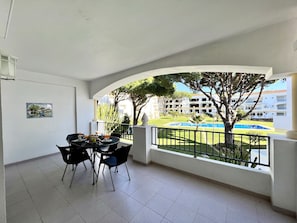 A balcony with a table set for dining, a view of a swimming pool, and a building in the background.