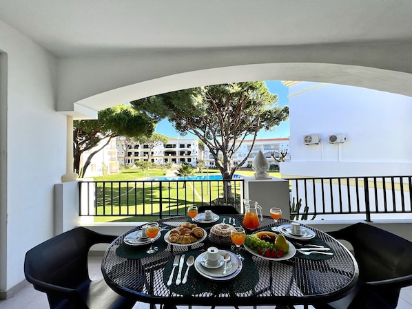 A balcony with a table set for breakfast, overlooking a pool and a building.