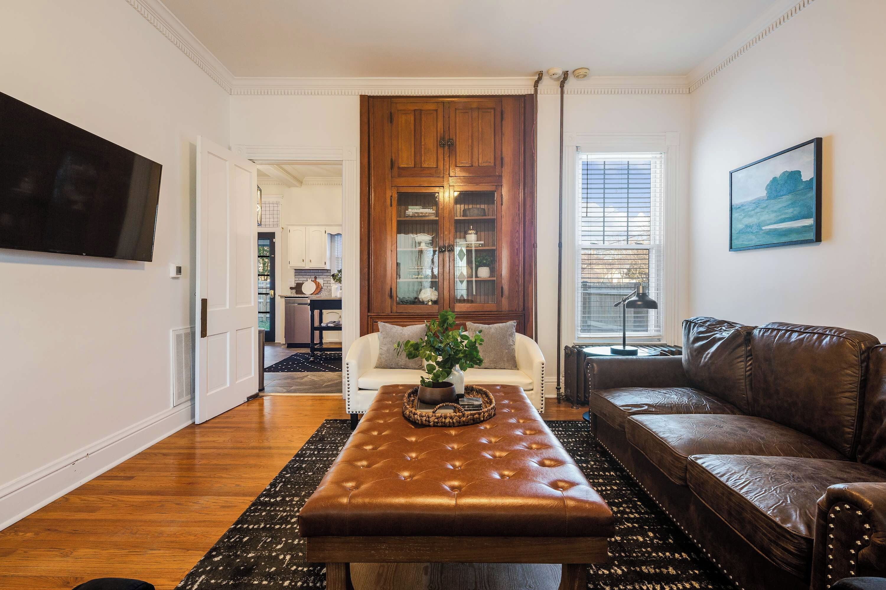 A living room with a brown leather ottoman, a white sofa, a brown leather couch, a wooden cabinet, and a painting on the wall.
