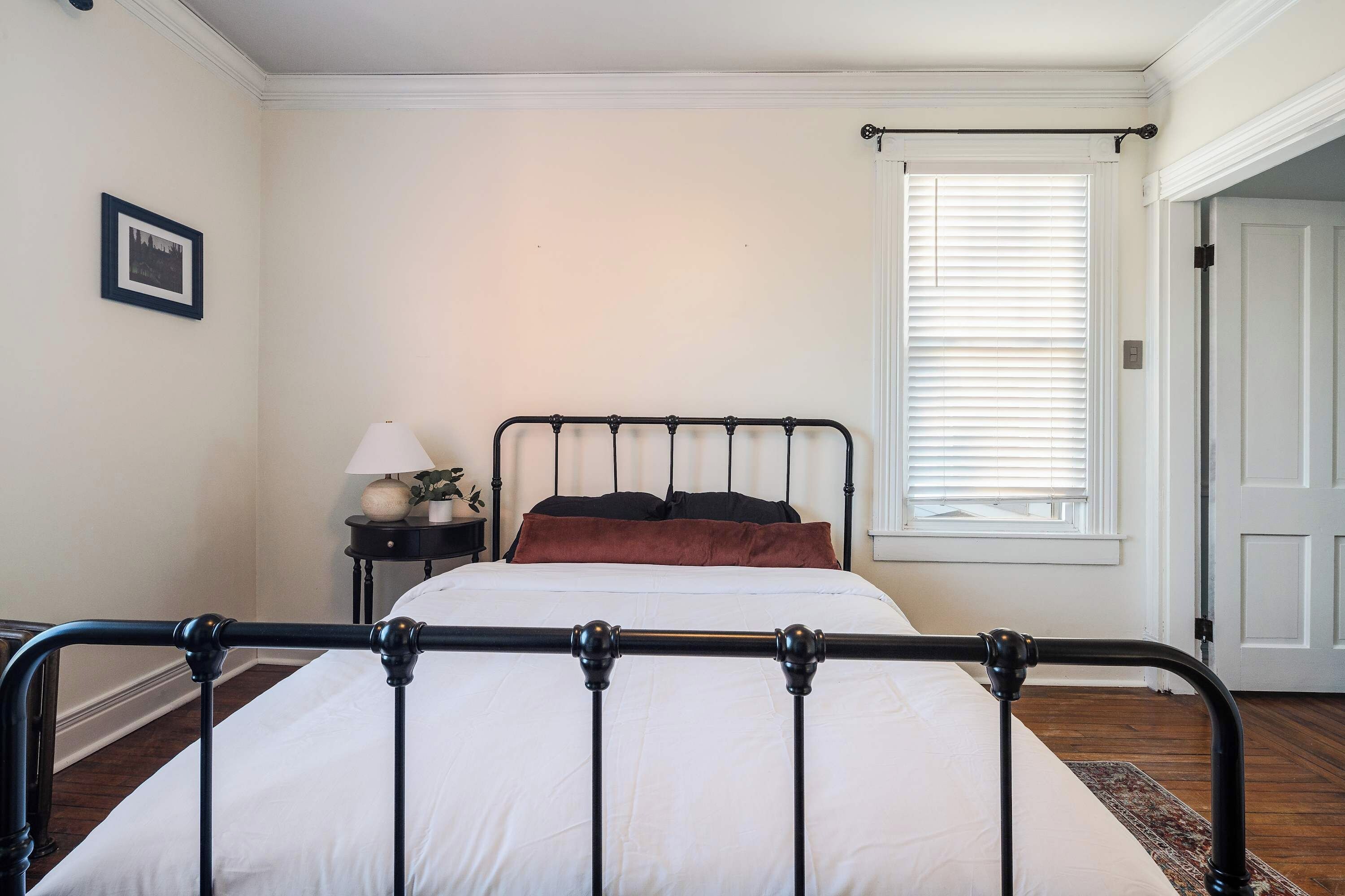 Quarto com cama, mesa de cabeceira, abajur, janela com persianas e porta.