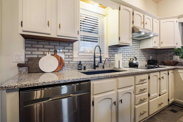 A kitchen with white cabinets, a granite countertop, a stainless steel dishwasher, and a window with blinds.