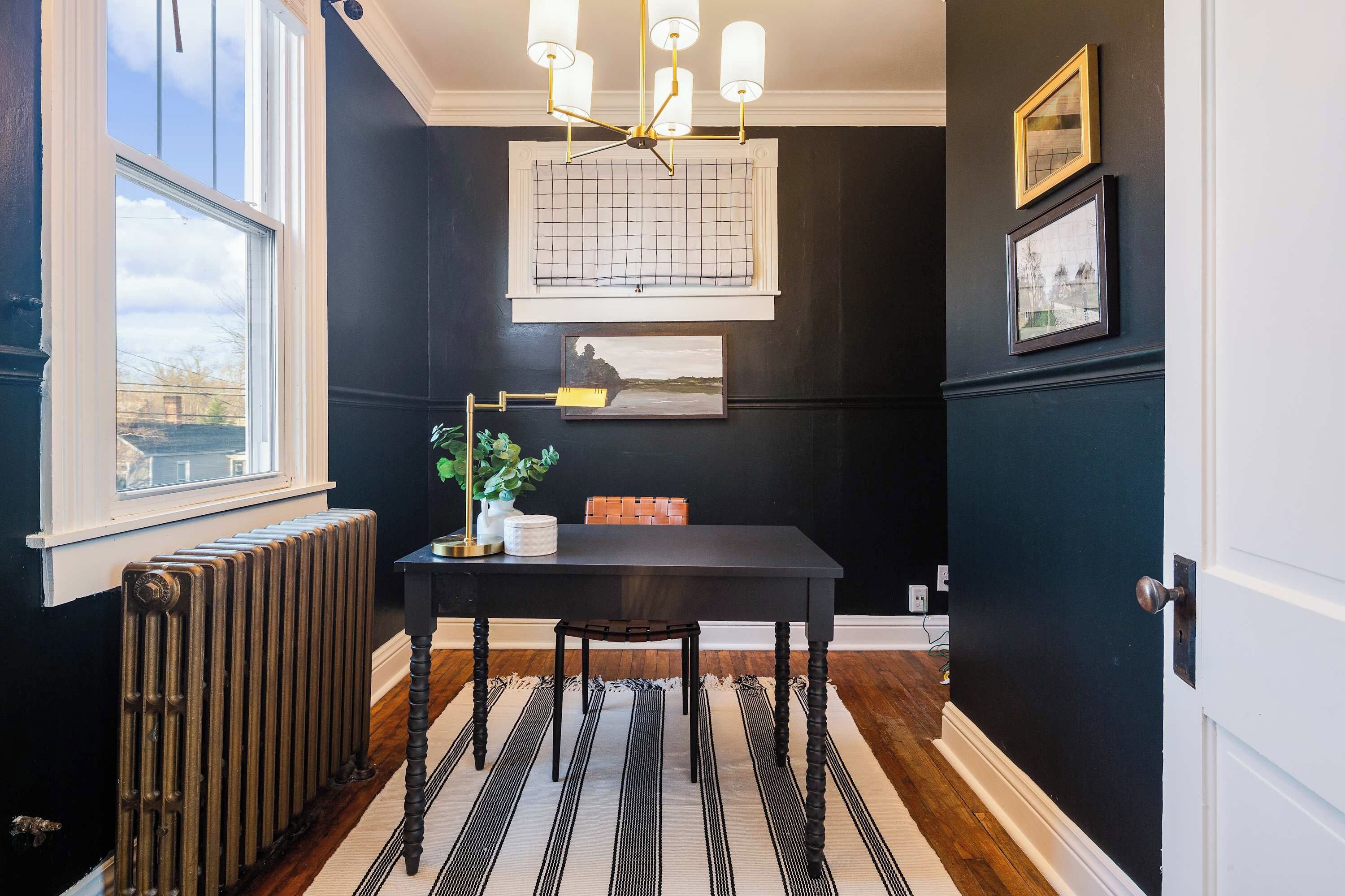 Een kamer met een zwarte muur, een raam, een radiator, een tafel met stoelen, een plant en kunstwerken.