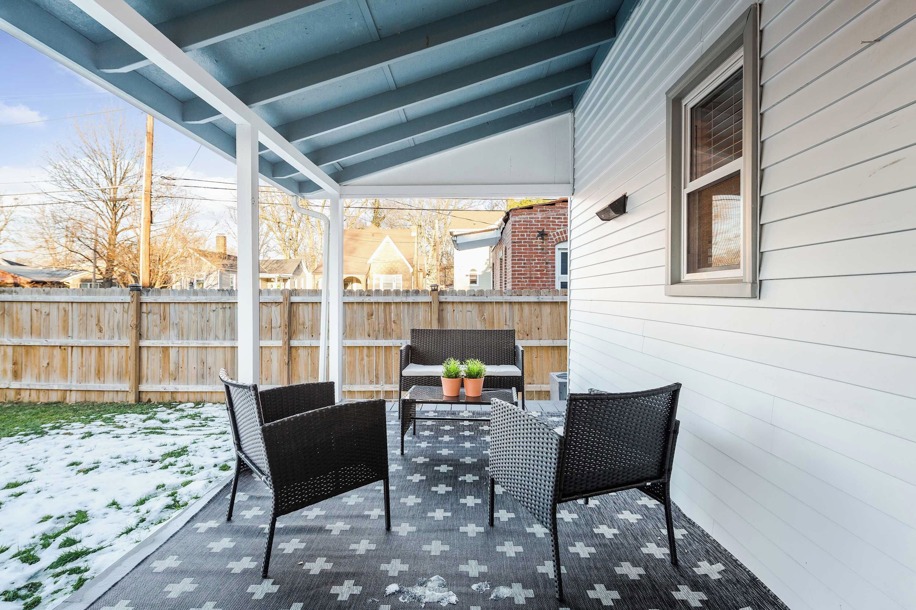 Eine überdachte Terrasse mit zwei Stühlen und einem Tisch, ein Holzzaun im Hintergrund und ein Haus mit Profilschalung.