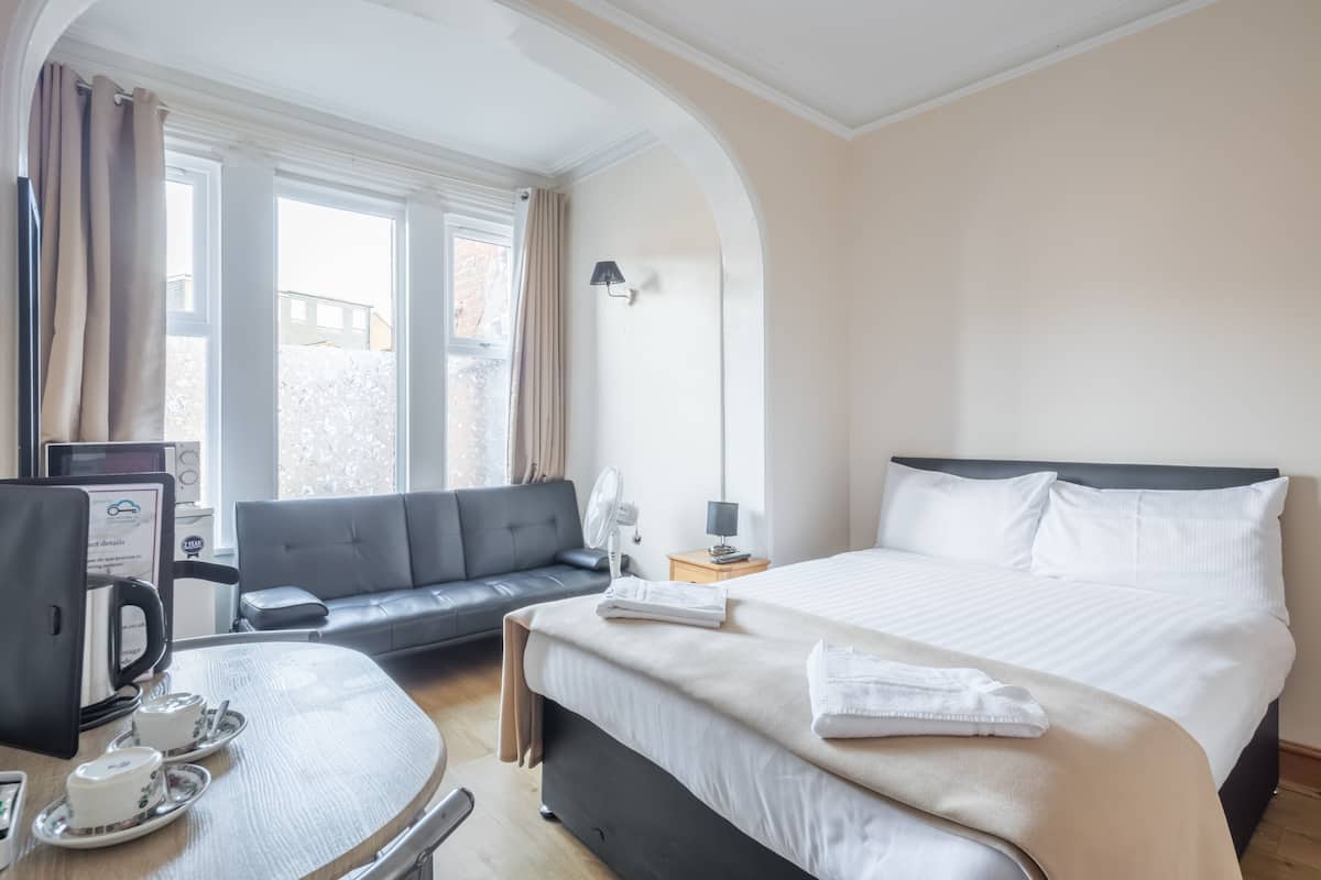 Une chambre d’hôtel avec un lit, un canapé, une table avec une théière et des tasses, un bureau avec un ordinateur et une fenêtre avec des rideaux.