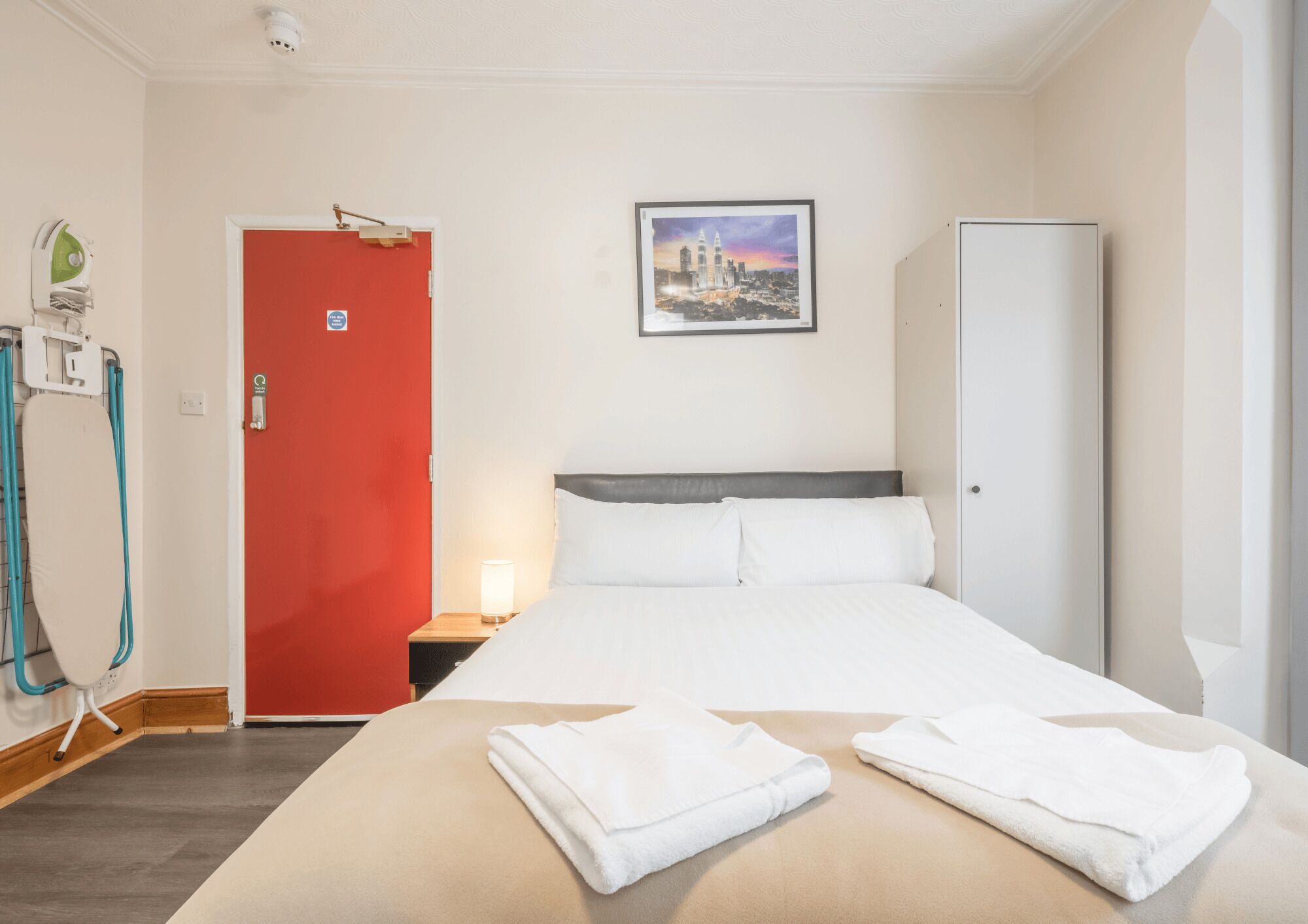Ein Hotelzimmer mit einem Bett, einer roten Tür, einem weißen Schrank, einem Nachttisch mit Lampe und einem Wandbild.
