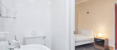 A modern bathroom with a white sink, toilet, and shower. There is a bed with a nightstand and a lamp in the adjacent room.