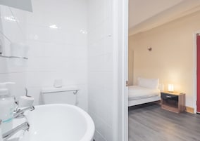 A modern bathroom with a white sink, toilet, and shower. There is a bed with a nightstand and a lamp in the adjacent room.