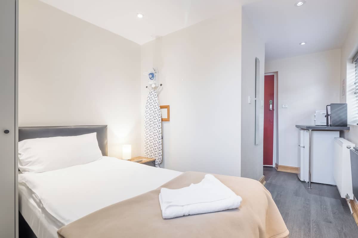 Une chambre d’hôtel avec un lit, une table de chevet avec une lampe, un petit bureau avec un ordinateur et une porte rouge donnant accès à la salle de bain.