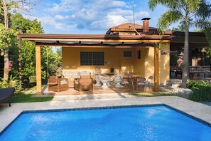 Et poolområde med en overdækket terrasse, udendørs møbler og en klar, blå pool.