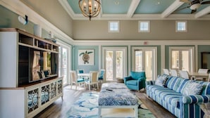 A spacious living area with a large sofa, a coffee table, and a TV on a cabinet. There are two doors leading to outdoor areas, and a ceiling fan is visible. - Cinnamon Beach Sunrise: Unit 853 (Palm Coast)