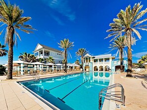 Une piscine entourée de chaises longues et de palmiers, elle-même bordée d’un hôtel composé de plusieurs bâtiments dotés de balcons.
