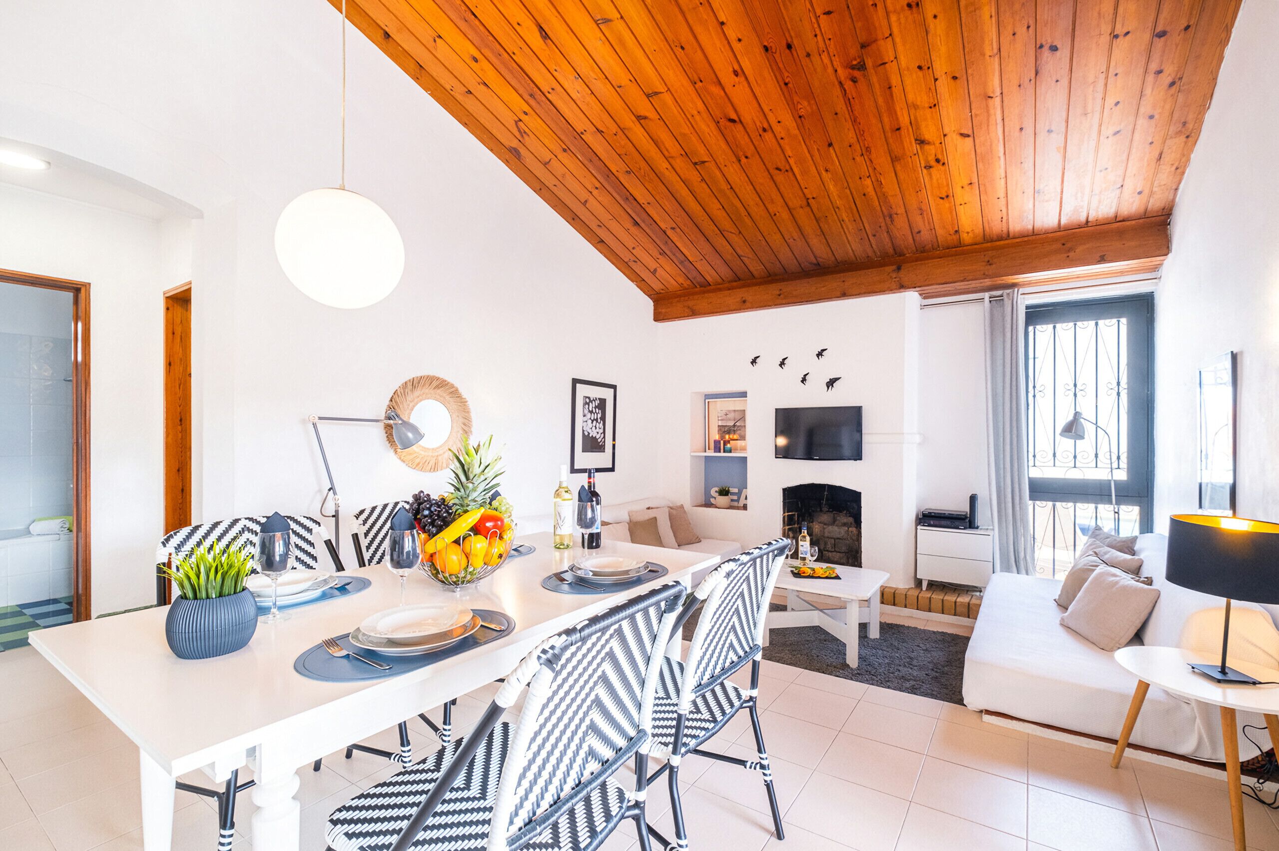 Un salon moderne avec un plafond en bois, un coin repas avec une table blanche et des chaises noires et blanches, un canapé et une cheminée.