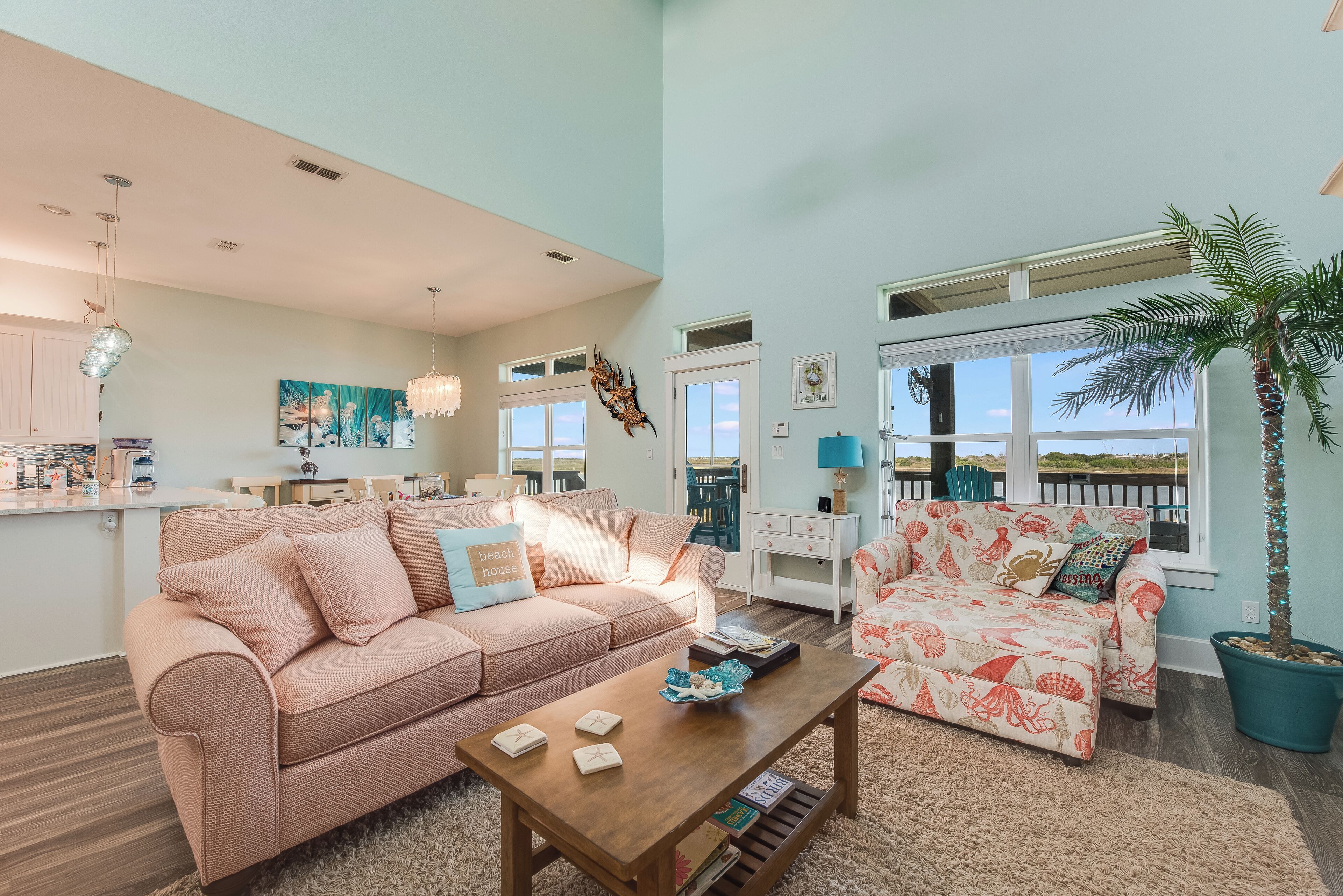 A spacious living room with a pink sofa, a patterned armchair, and a wooden coffee table. There is a large window with a view of the outdoors.