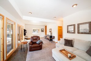 A spacious living room with a sofa, a leather chair, and a coffee table. There are framed pictures on the wall and a desk with a chair in the background.