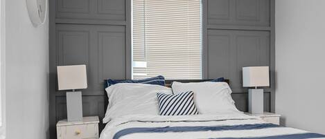Un dormitorio con una cama, dos mesitas de noche con lámparas, una ventana con persianas y un reloj de pared.