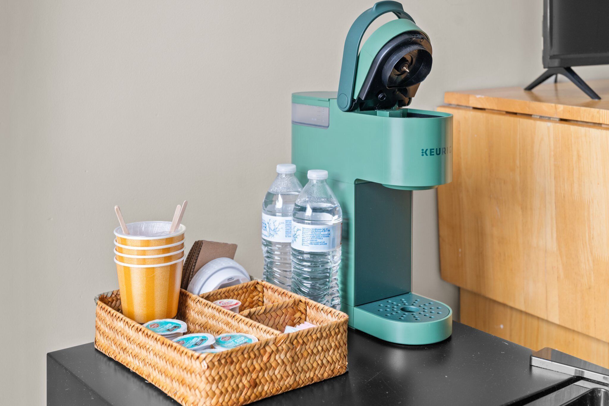 Una cafetera color turquesa, dos botellas de agua, una cesta de mimbre con una taza y un recipiente, y una encimera negra.