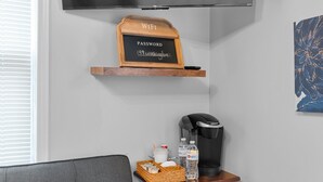 A modern living room with a flat-screen TV mounted on the wall, a wooden shelf with a chalkboard displaying a WiFi password, a coffee maker, a basket with breakfast items, and a water bottle. - Washington Park Motel (Norwell)