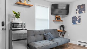 A modern living room with a grey sectional sofa, a built-in microwave, a small table with a plant, and a flat-screen TV mounted on the wall. - Washington Park Motel (Norwell)
