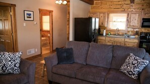Un salon avec un canapé gris et des coussins à motifs, une porte en bois et une cuisine avec des meubles en bois et un réfrigérateur noir.