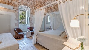 A bedroom with a brick ceiling, a bed with white bedding, a chair, a bedside table, and a window with a view of greenery. - Masseria Profilo 8, Emma Villas (Monopoli)