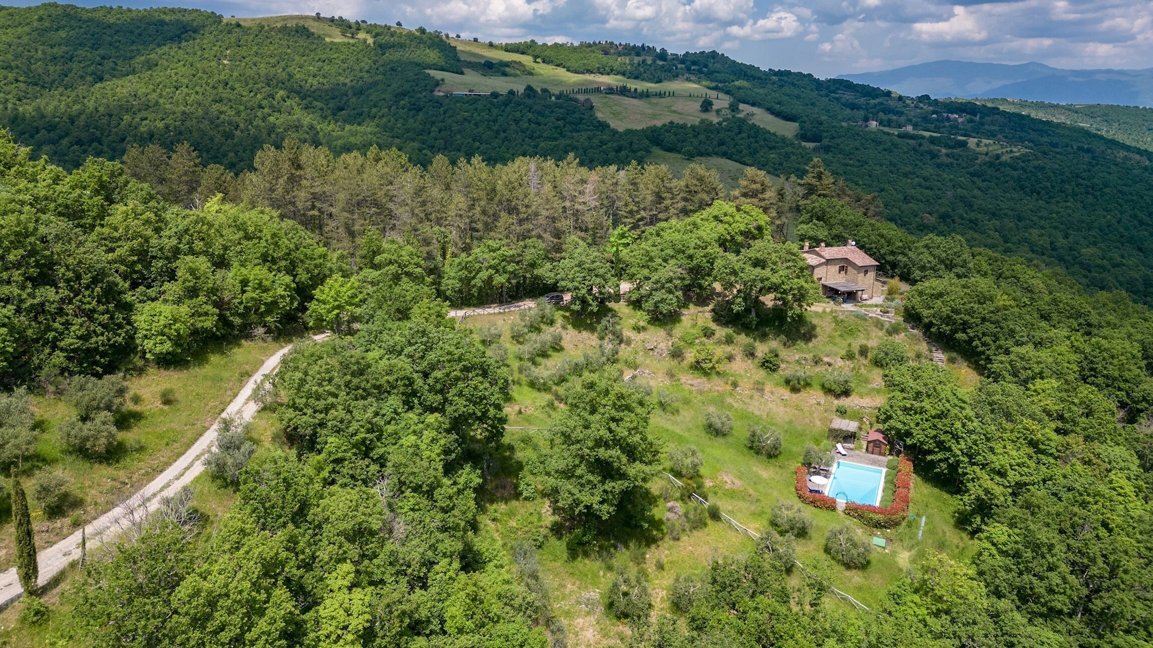 Una propiedad rural con una casa de piedra rodeada de bosque denso, un camino de tierra y una piscina.