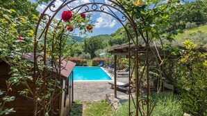 Ein Poolbereich mit einer Pergola, die mit Kletterrosen bewachsen ist, einer Holzkonstruktion und Blick auf ein Gebirge.