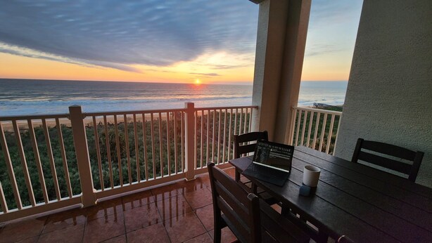 Ein Balkon mit Blick auf den Sonnenuntergang über dem Meer, ein Laptop und eine Tasse auf dem Tisch.