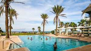 Ein Schwimmbad mit Palmen und Liegestühlen, umgeben von einer Poollandschaft mit Blick aufs Meer.