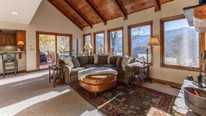 A spacious living room with a large sectional sofa, a wooden ceiling, and large windows offering mountain views.