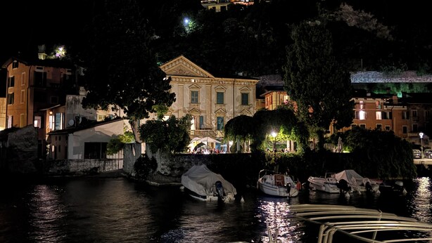 A historic building with classical architecture, situated by a calm body of water with boats docked nearby.
