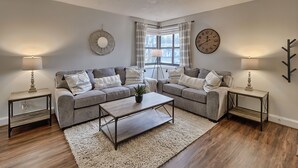 Ein Wohnzimmer mit zwei grauen Sofas, einem Holztisch und zwei Beistelltischen mit Lampen. Es gibt ein großes Fenster mit Vorhängen und eine Wanduhr.