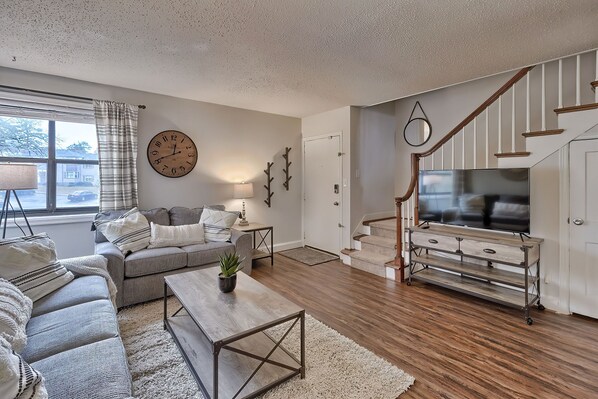 Relax in this stylish living room with a sofa, coffee table, and TV on a wooden stand, surrounded by hardwood flooring and a large wall clock.