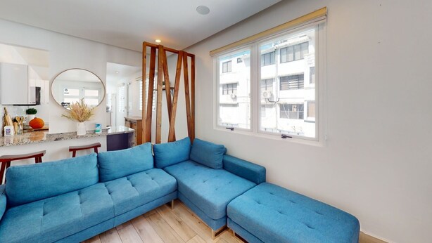 A modern living room with a blue sectional sofa, a wooden accent wall, a large window with a view of buildings, and a kitchen area with a countertop and sink. - Beautiful Gem In The Heart of Condado - 3 mins from the beach! (San Juan)