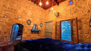 A rustic stone-walled room with a bed, a small window with a view, a wall-mounted fan, and a decorative mirror. - Ultimate experience of living in a traditional Legacy with a natural setting (Sidon)