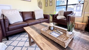 Ein Wohnzimmer mit braunem Ledersofa, einem Holztisch und einem großen Fenster mit Blick auf Gebäude.