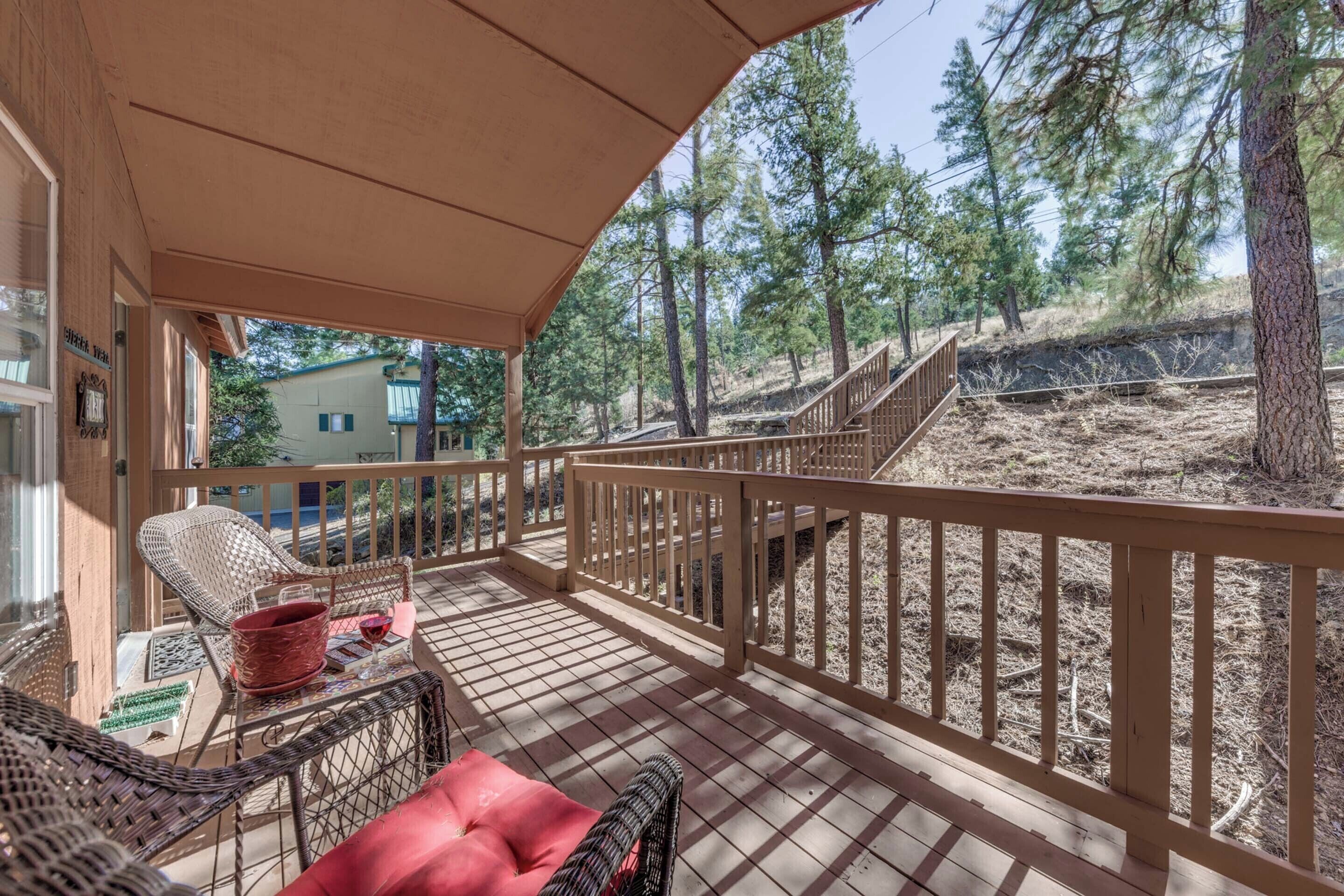 A wooden deck with wicker furniture and a red cushion, overlooking a forested area with a staircase.
