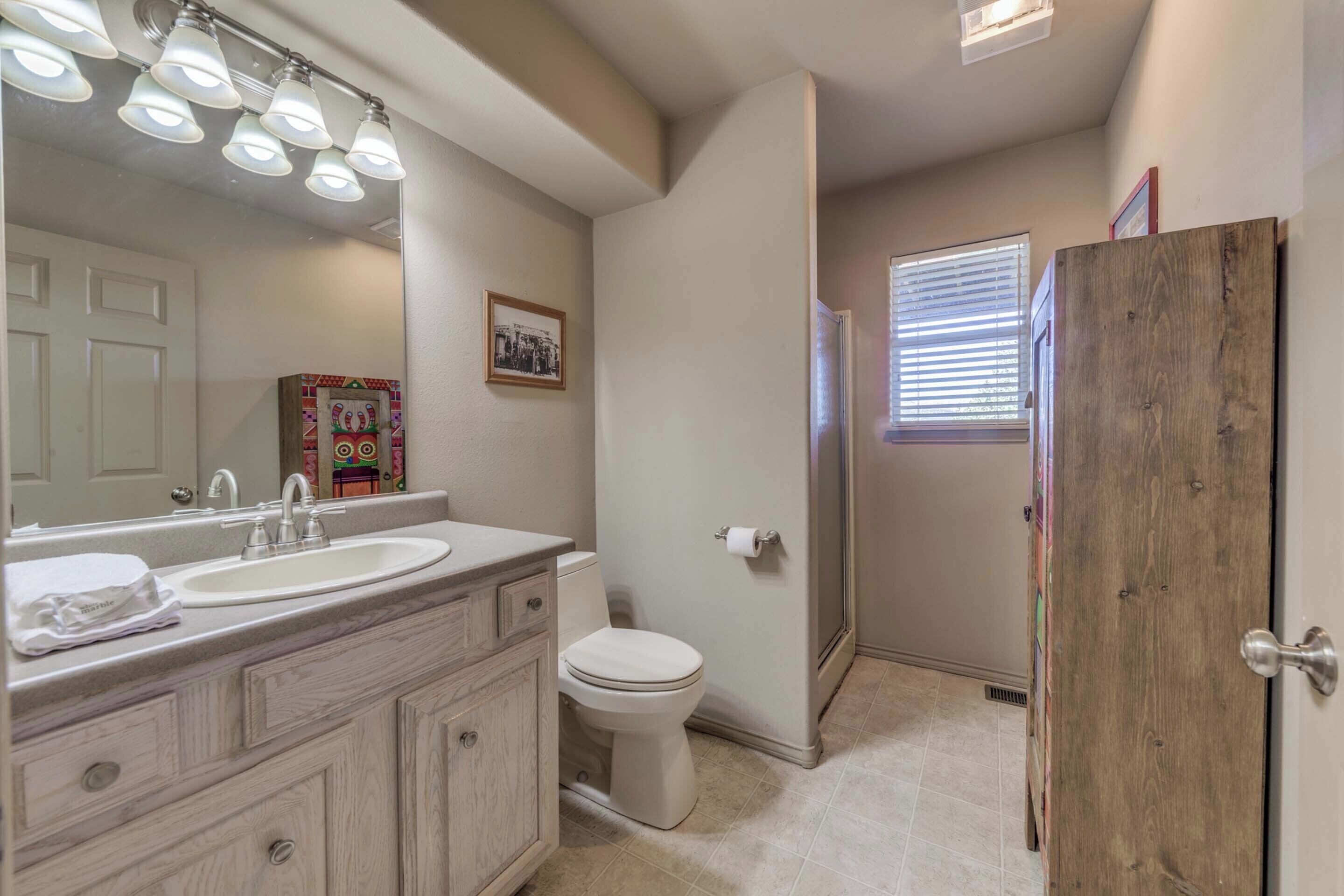 A bathroom with a sink, mirror, and toilet. There is a wooden door and a window with blinds.