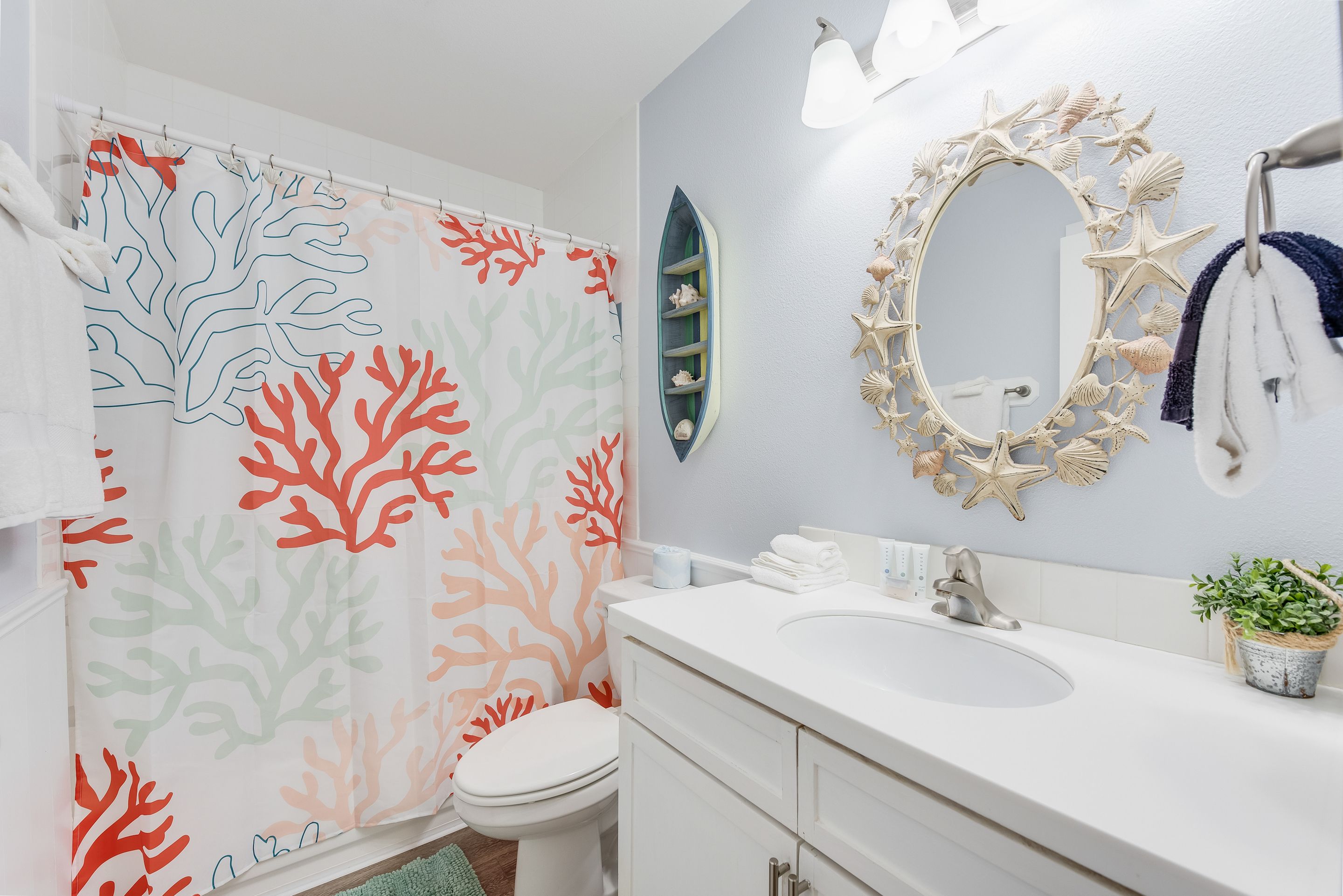 A bathroom with a coral-patterned shower curtain, a white sink, a mirror with a starfish design, and a small plant on the counter.