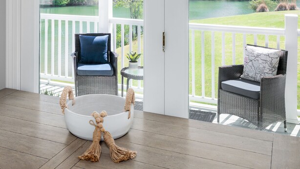 A spacious room with a large sliding glass door leading to a deck. Inside, there is a round white table with a decorative item, two wicker chairs with cushions, and a small round table with a plant.