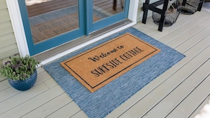 A blue and brown doormat with "Welcome to SURFSIDE COTTAGE" text, a potted plant, and a black table with baskets underneath a door.
