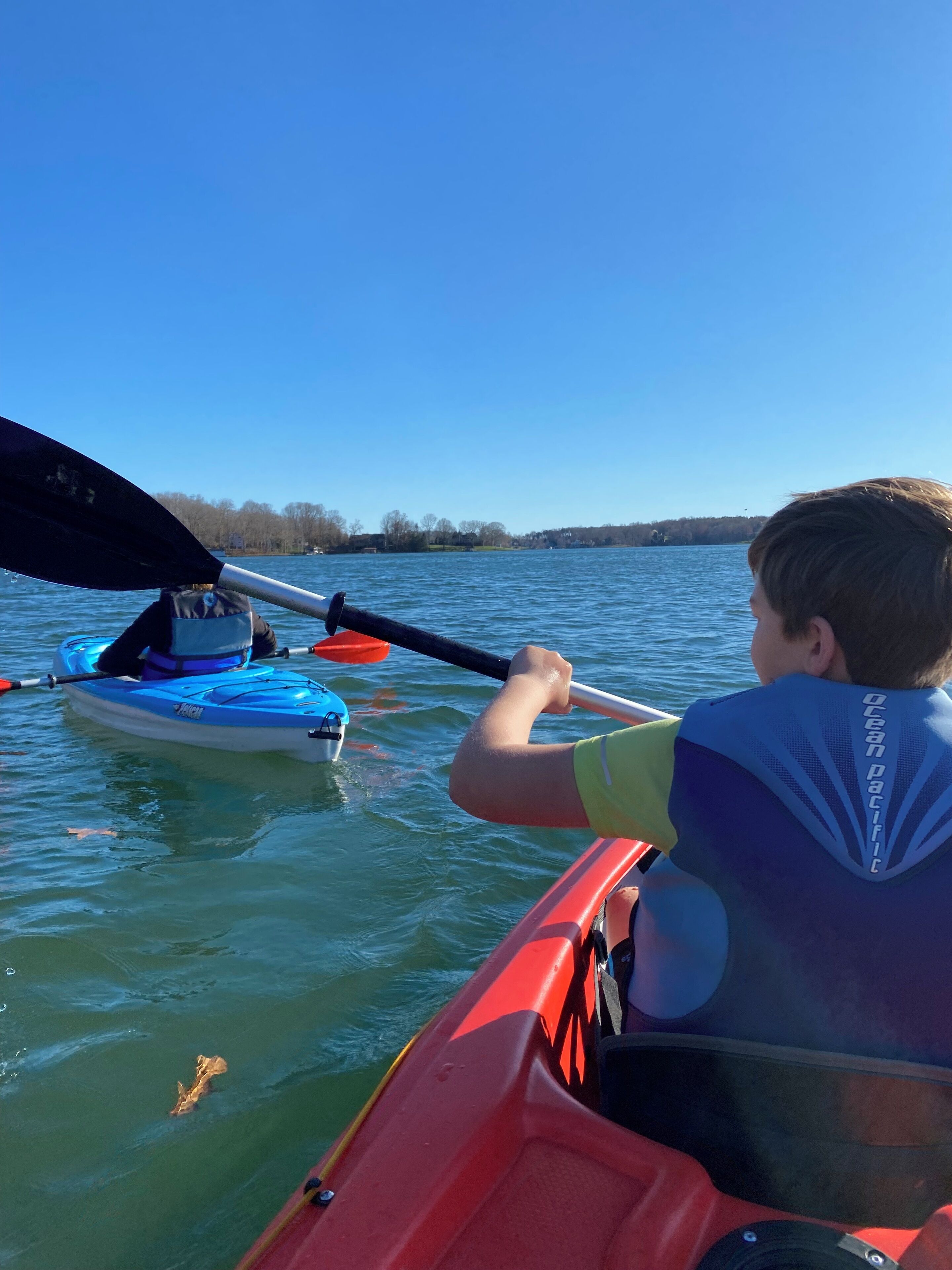 Une personne dans un kayak rouge pagaye sur un lac calme, avec un kayak bleu en arrière-plan.