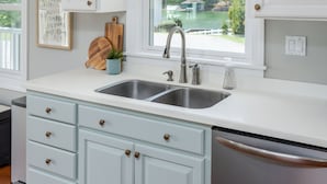 A kitchen with white cabinets, a double sink, a window with a view of greenery, and a pendant light.