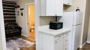 A compact kitchen with white cabinets, a microwave, and a refrigerator. There is a wooden floor and a rug in the living area. - Scout- a rustic “cabin” apartment, very fast wifi (Salem)