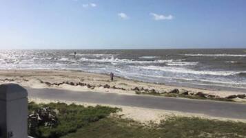 Une vue sur la plage, avec une route parallèle au rivage, une personne qui marche et un ciel dégagé.