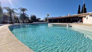 A clear swimming pool with a tiled edge, surrounded by palm trees and a building with a covered patio area. - São Rafael Beach House (Albufeira)