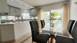 A modern kitchen with a central island, dining area with glass table and chairs, and a view of greenery outside. - São Rafael Beach House (Albufeira)